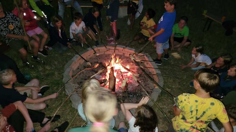 Eine Gruppe von Kindern sitzt um ein Lagerfeuer und hält Knüppel mit Stockbrot über die Flammen.